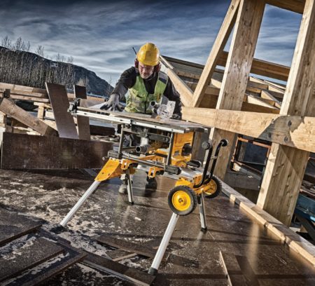 A construction worker in a yellow hard hat cuts wood on a table saw surrounded by building materials.