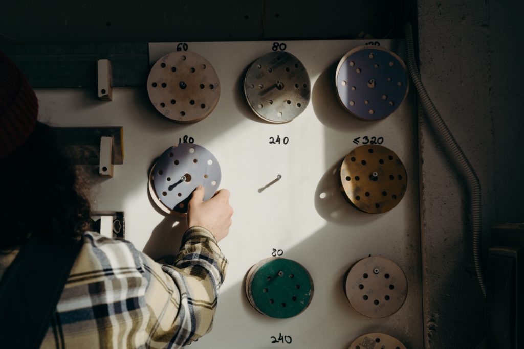 A person in a brown and black plaid shirt chooses a sanding disc from several hanging on a wall.