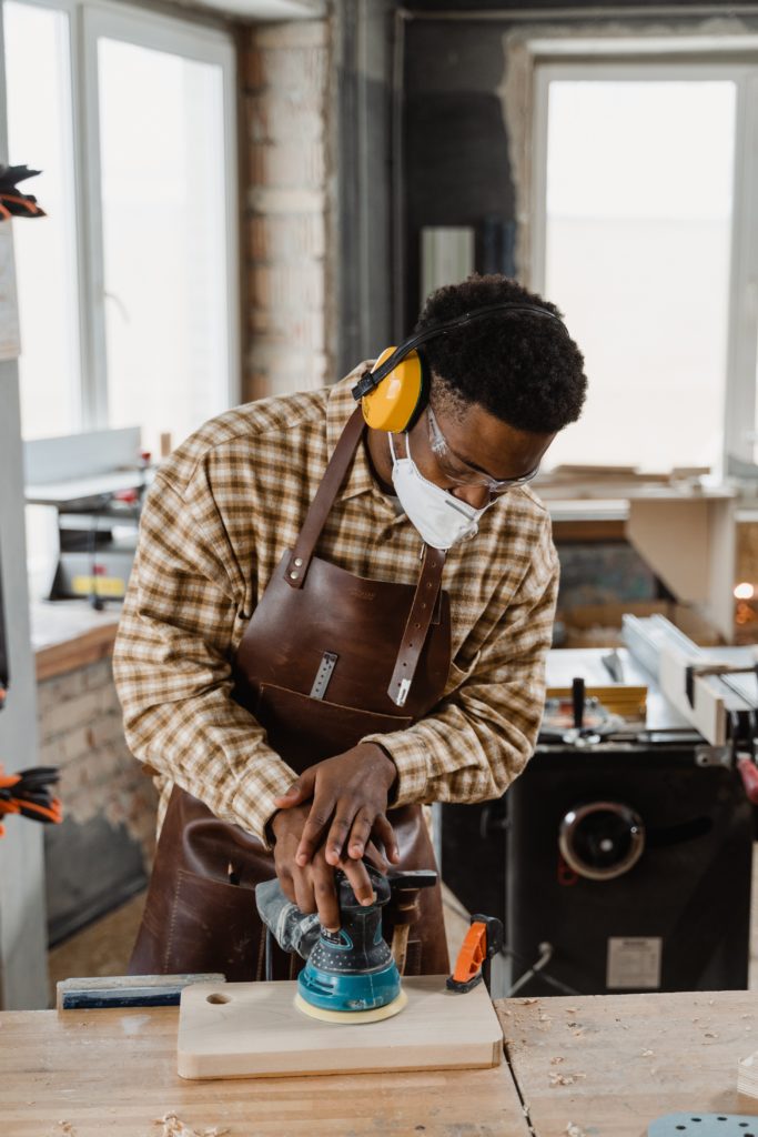 A man wearing a mask and earmuffs uses an orbital sander on a piece of wood.