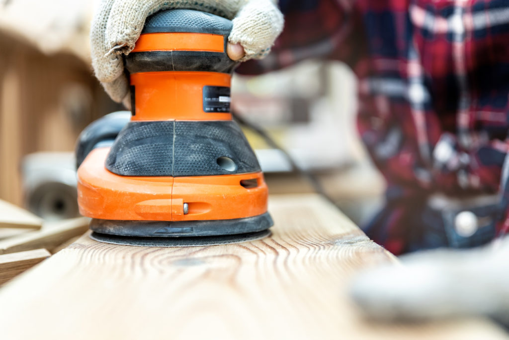 A handheld orbital sander grinding against a raw wood plank