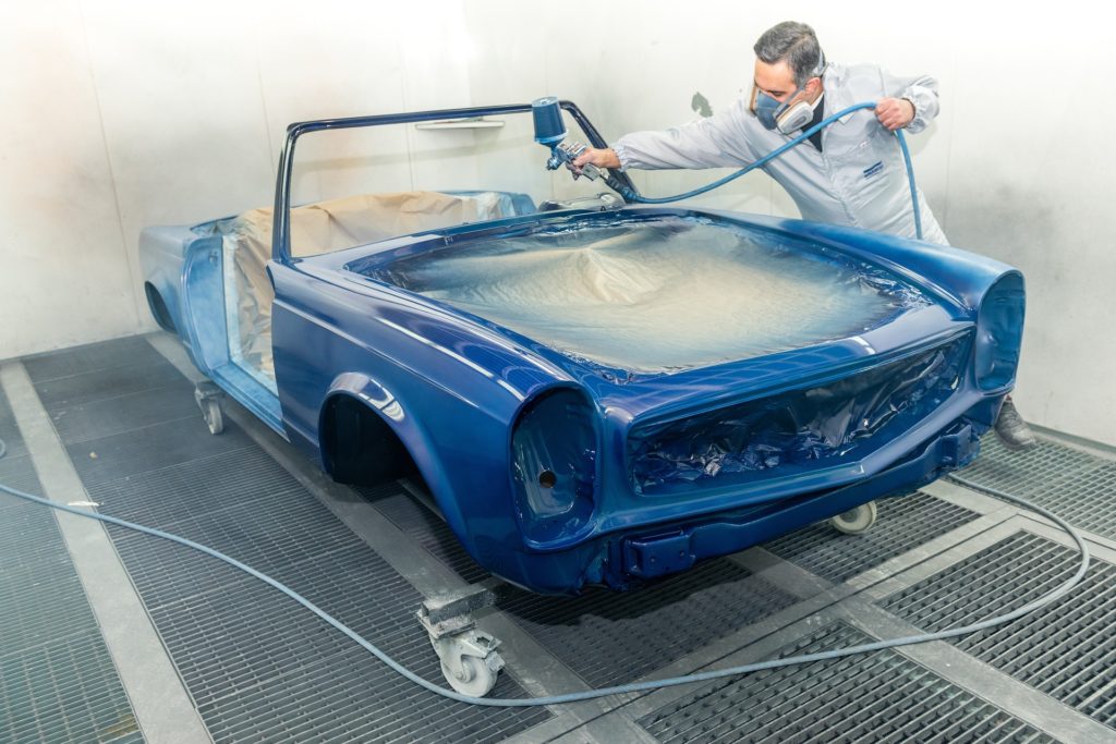 A man in a painting booth uses an HVLP gun to paint a bright blue classic convertible.