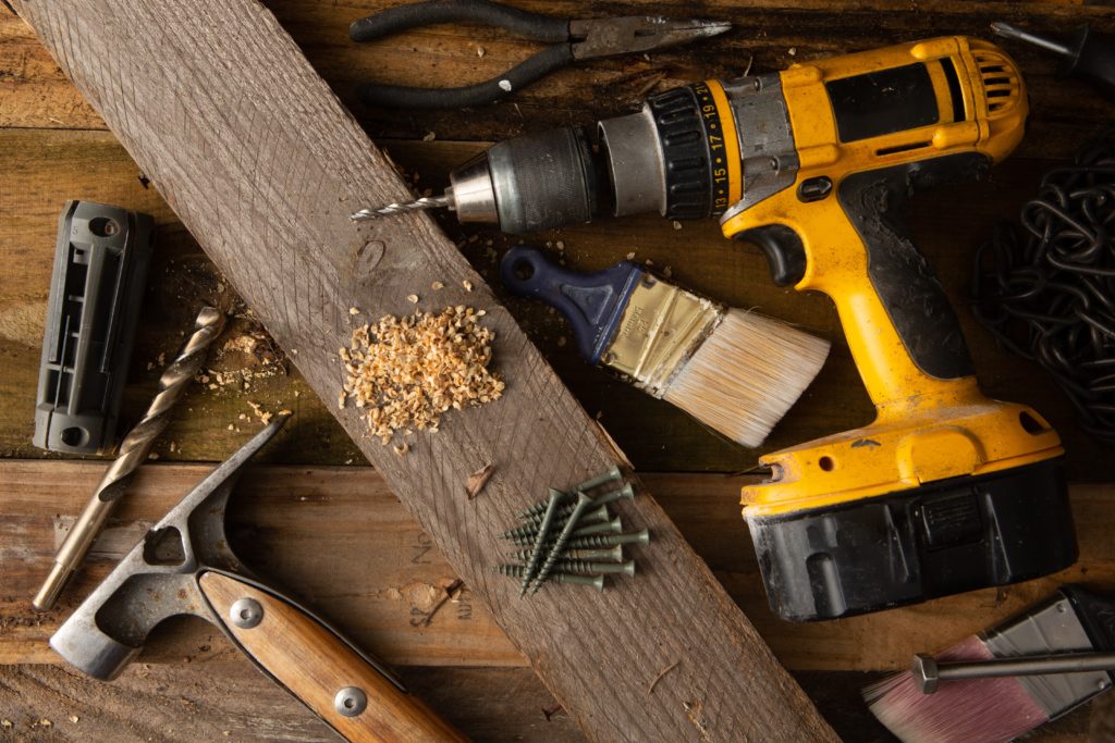 A variety of tools, including a hammer and cordless drill, sit on top of a table made of wood boards.