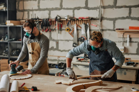 Warm toned portrait of two carpenters in protective gear working with wood while building handicraft furniture in workshop.