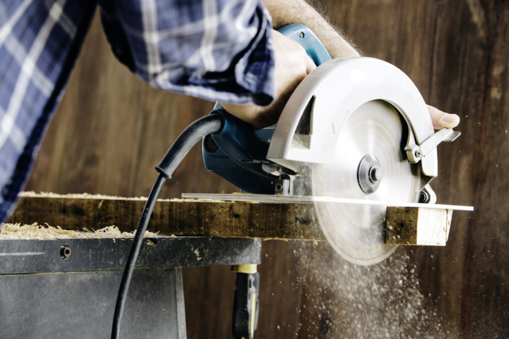 Male carpenter using electric circular saw in home workshop with wood chips flying.