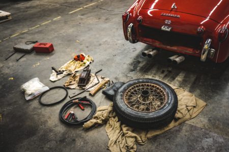 A floor jack, jumper cables, and other tools in a shop with a classic red Triumph automobile.