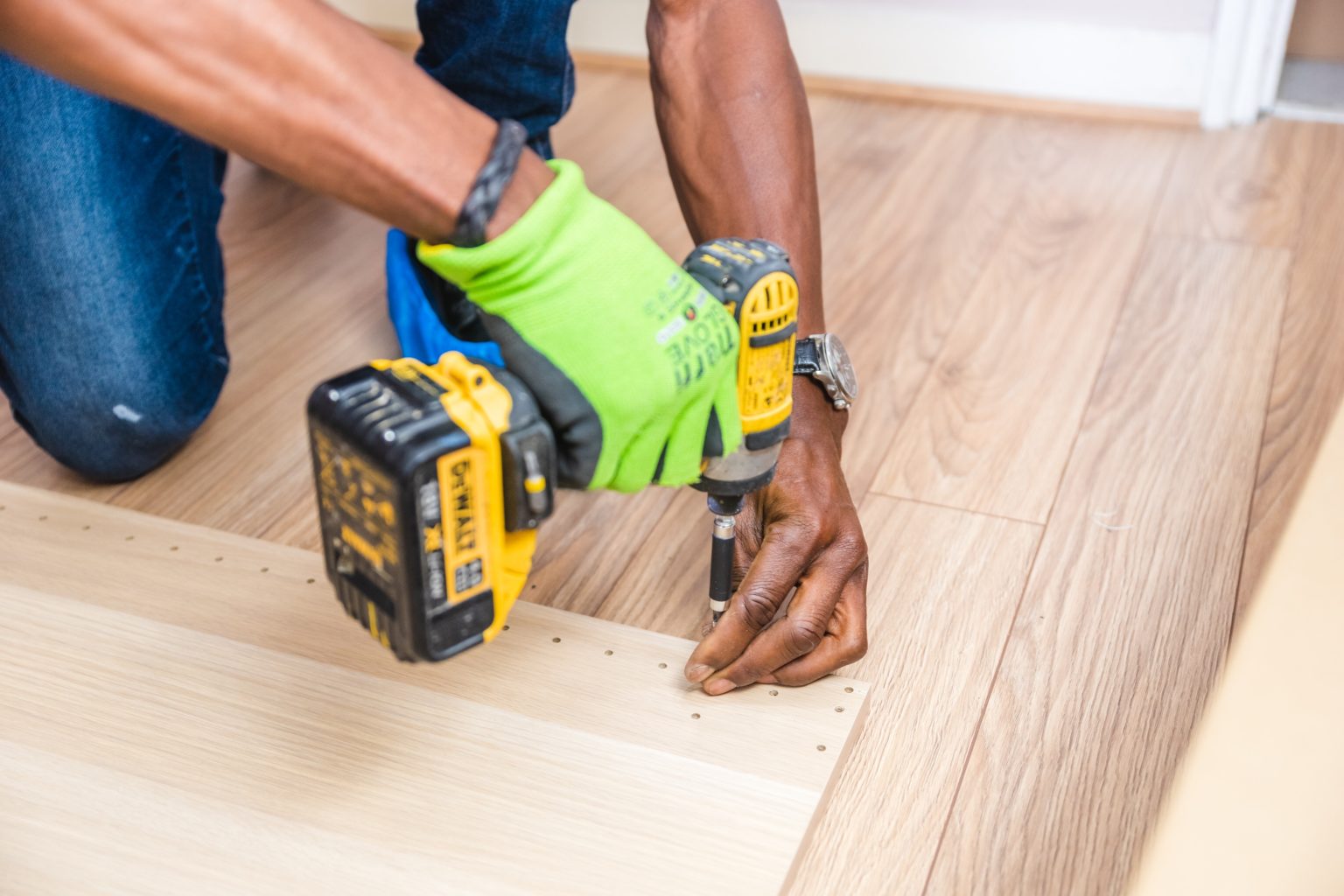 A person with medium brown skin and green gloves uses a yellow drill to put holes in wood.