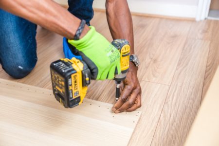 A person with medium brown skin and green gloves uses a yellow drill to put holes in wood.