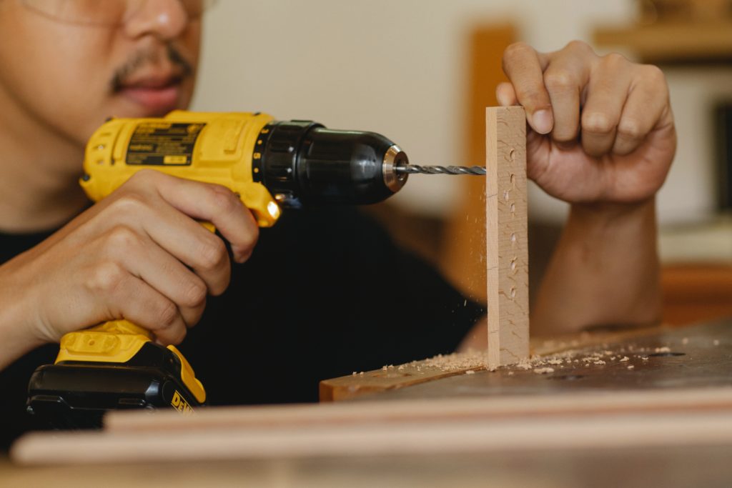 A person uses a yellow power drill to put a row of holes in a small piece of wood.
